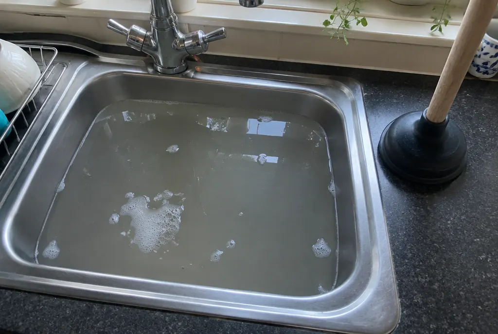Stainless steel kitchen sink full of water that won't drain, with a plunger beside it