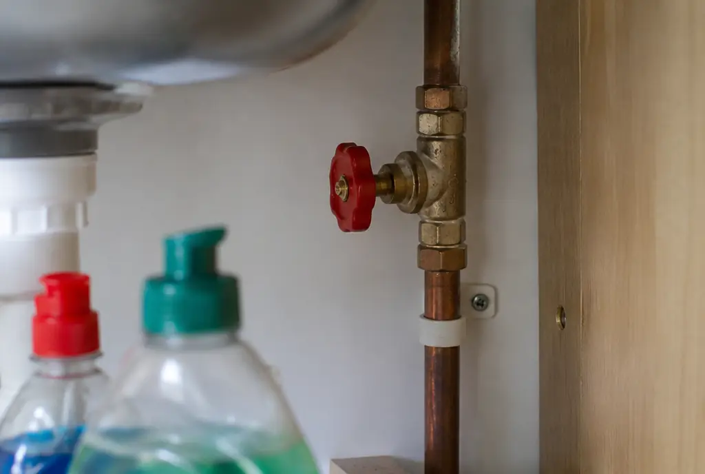 Brass stopcock with a red handle on a copper pipe under a kitchen sink
